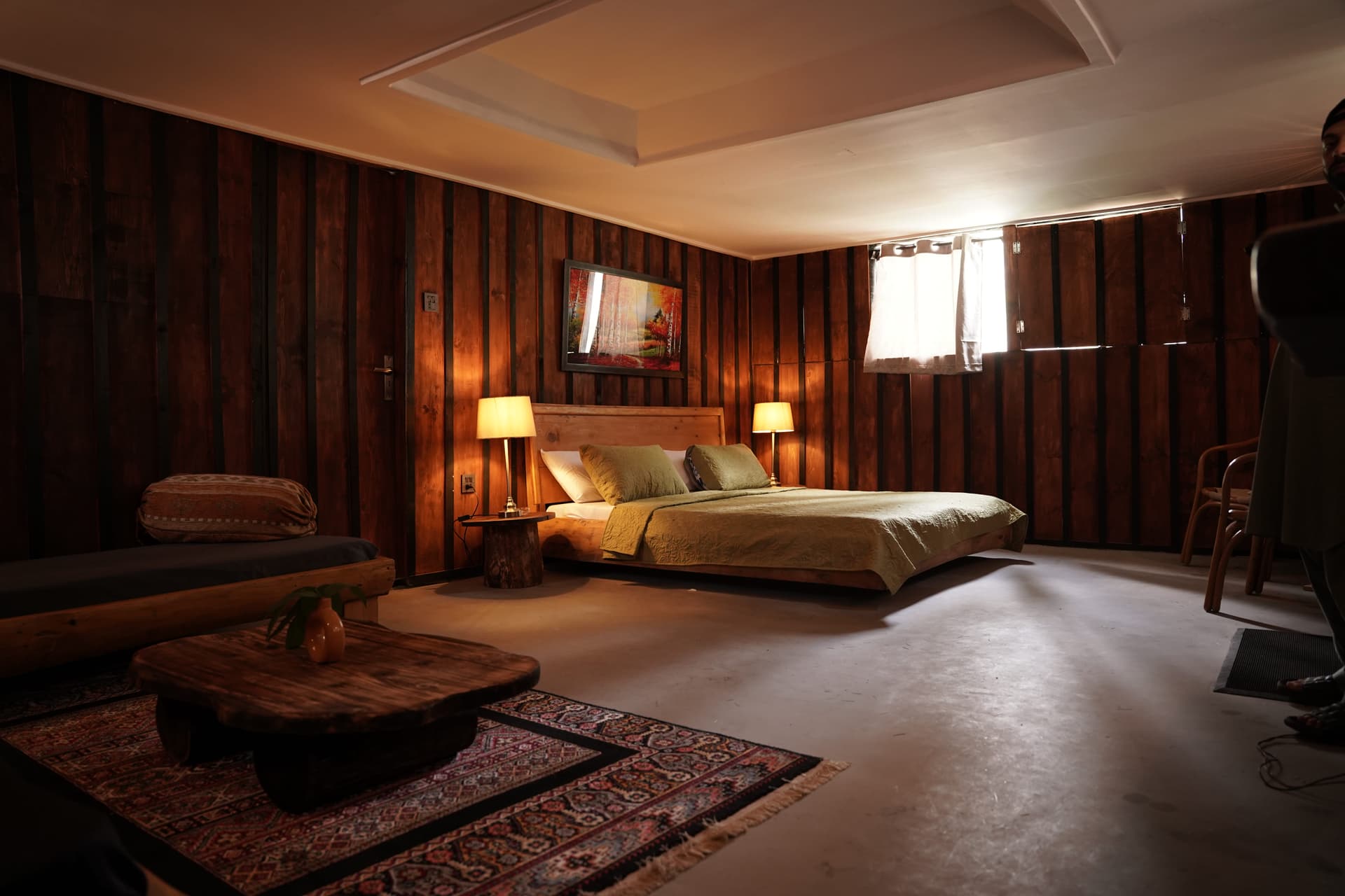 Spacious bedroom with warm wood paneling, olive green bedding, rustic furniture, and patterned rug