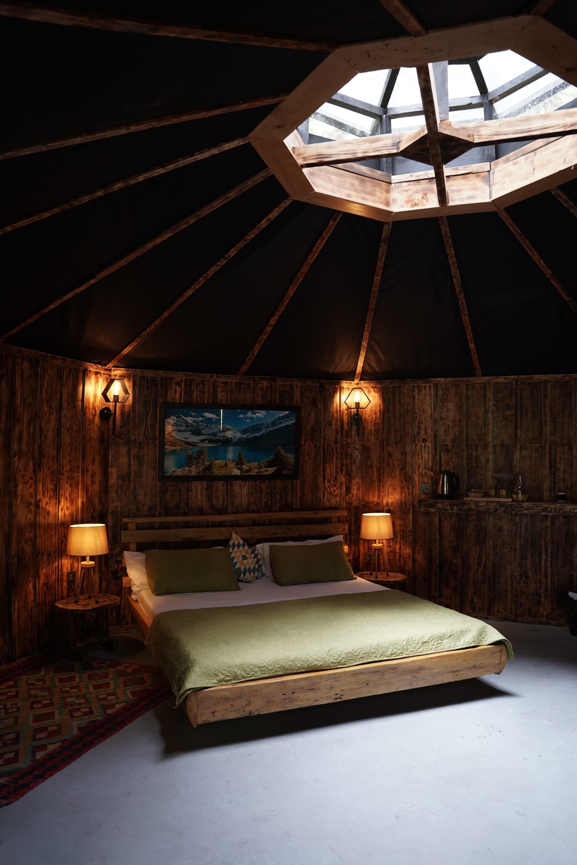 Yurt-style bedroom with octagonal skylight, dark wood walls, sage green bedding, and dramatic ceiling beams